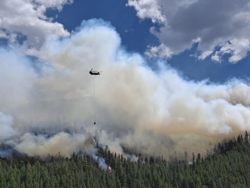 A fire-fighting helicopter flies over burning trees and a sky full of smoke.