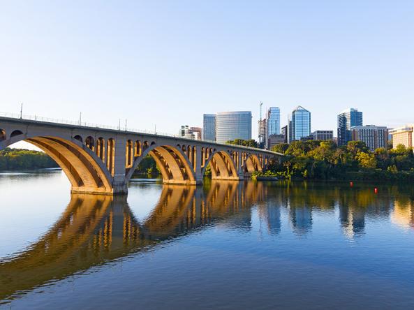 Key Bridge and the Rosslyn skyline in Arlington, Virginia, overlooking the Potomac River. The concrete arch bridge spans from the left foreground toward the right, with its golden-hued arches reflected clearly in the calm blue water below. In the background, a cluster of modern high-rise office and residential buildings sits atop a wooded shoreline under a clear, pale blue sky.