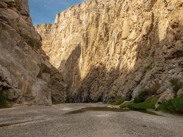  A low-angle view of nearly dry section of the Rio Grande as it flows through the narrow cliffs of Santa Elena Canyon in West Texas. Towering, steep limestone walls rise on both sides. The canyon floor is dominated by a wide expanse of dry grey gravel and silt, with only a small, shallow pool of green water remaining on the right side. Sparse green vegetation and reeds cling to the base of the canyon walls where moisture is still present.