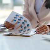 A woman in a blazer looks through a clipboard of charts and graphs at a desk. Photo credit: amnaj, Adobe Stock.