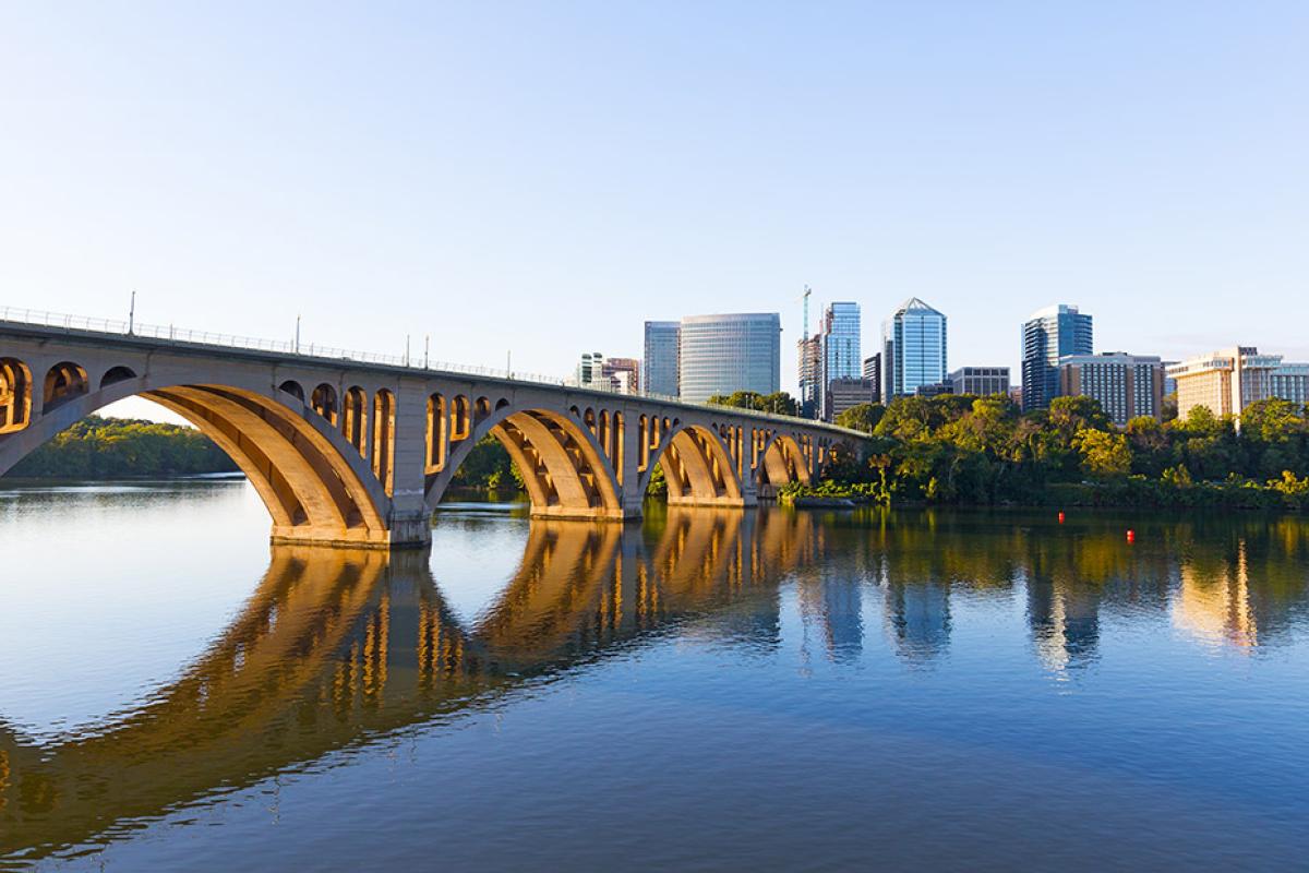 Key Bridge and the Rosslyn skyline in Arlington, Virginia, overlooking the Potomac River. The concrete arch bridge spans from the left foreground toward the right, with its golden-hued arches reflected clearly in the calm blue water below. In the background, a cluster of modern high-rise office and residential buildings sits atop a wooded shoreline under a clear, pale blue sky.