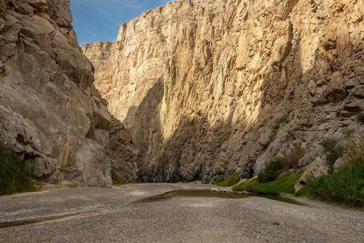  A low-angle view of nearly dry section of the Rio Grande as it flows through the narrow cliffs of Santa Elena Canyon in West Texas. Towering, steep limestone walls rise on both sides. The canyon floor is dominated by a wide expanse of dry grey gravel and silt, with only a small, shallow pool of green water remaining on the right side. Sparse green vegetation and reeds cling to the base of the canyon walls where moisture is still present.